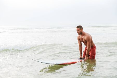 Image of a concentrated strong young handsome man surfer with surfing on a beach outside.の写真素材