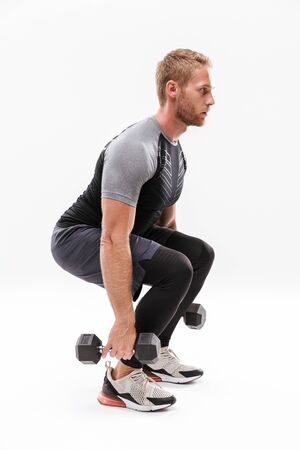 Confident attractive young muscular sportsman standing isolated over white background, lifting heavy barbells, squattingの写真素材