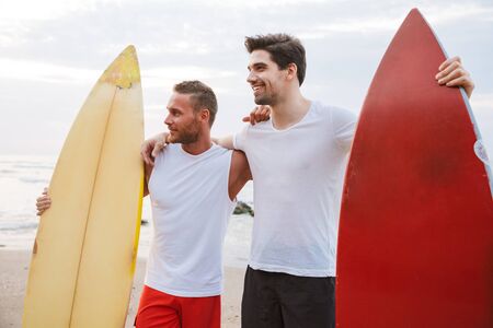 Image of a happy young cheery positive two men surfers friends with surfings on a beach outside.の写真素材