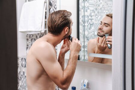 Attractive young shirtless man shaving while standing at the bathroom mirror, using electrical razorの写真素材