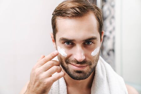 Attractive young shirtless man applying facial cream while standing at the bathroom mirrorの写真素材