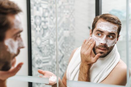 Attractive young shirtless man applying facial cream while standing at the bathroom mirrorの写真素材