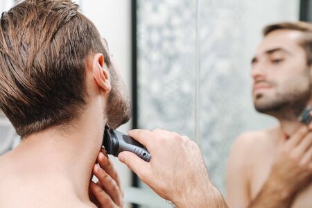 Attractive young shirtless man shaving while standing at the bathroom mirror, using electrical razorの写真素材