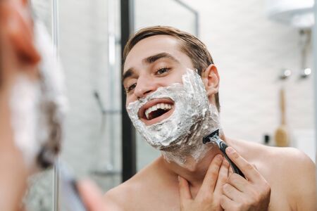 Attractive young shirtless man shaving while standing at the bathroom mirrorの写真素材