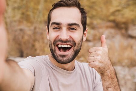 Image of unshaven laughing man showing thumb up and taking selfie photo while walking on sunny beachの写真素材