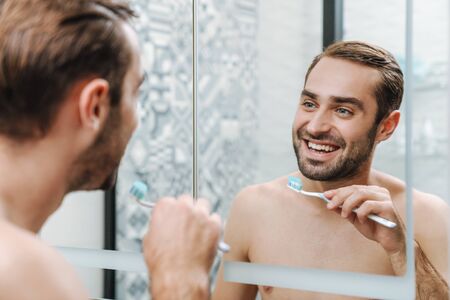 Attractive smiling shirtless man brushing teeth while standing at the bathroom mirrorの写真素材