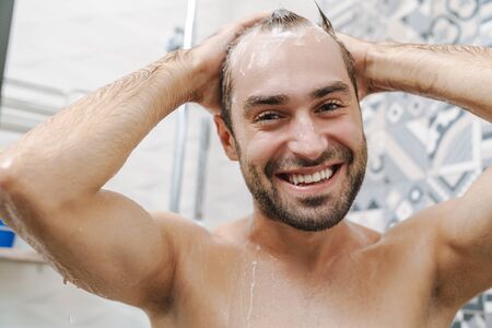 Attractive young smiling man washing hair while standing in the shower