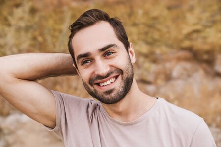 Image of unshaven caucasian man smiling and looking forward while walking on sunny beach