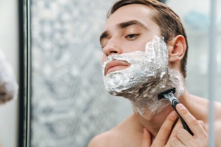Attractive young shirtless man shaving while standing at the bathroom mirrorの写真素材