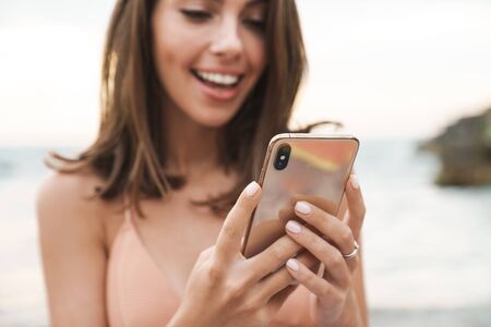 Image of nice cheerful woman in bra smiling and using cellphone while resting on sunny beachの写真素材