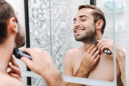 Attractive smiling young shirtless man shaving while standing at the bathroom mirror, using...
