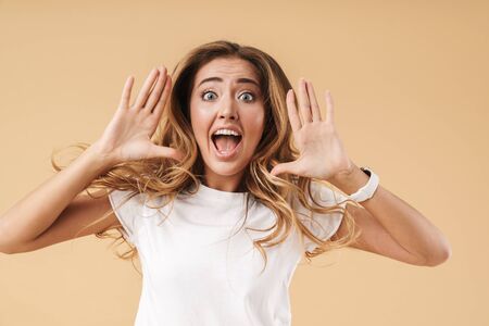 Portrait of an attractive excited young woman with long curly blonde hair wearing casual clothing standing isolated over beige background, screamingの写真素材