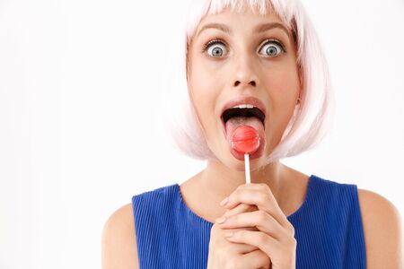Portrait of young surprised woman wearing pink wig licking lollipop at camera isolated over white backgroundの写真素材