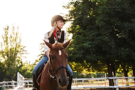 Image of happy young blonde woman wearing hat riding horse at yard in countrysideの写真素材