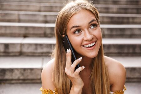 Beautiful smiling young girl talking on mobile phone while sitting on stairs outdoors