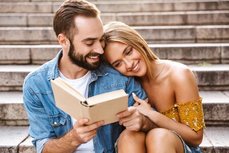 Smiling lovely young couple sitting together on stairs, reading a book, huggingの写真素材