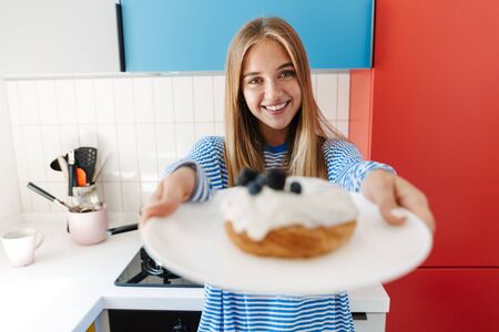 Image of a smiling cheery young cute girl at home at kitchen give you a sweet donut.