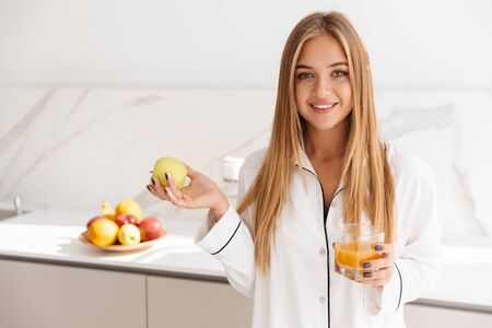 Photo of smiling attractive woman in pajama holding apple and drinking juice while standing in bright kitchenの写真素材