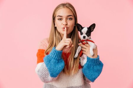 Cute lovely girl playing with her pet chihuahua isolated over pink background, showing silence gestureの写真素材