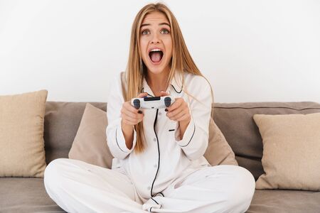 Photo of excited blonde woman in pajama playing video game while sitting on sofa in bright living...