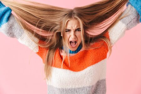 Portrait of an annoyed blonde girl wearing sweater screaming isolated over pink background, pulling her hair outの写真素材