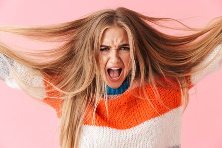 Portrait of an annoyed blonde girl wearing sweater screaming isolated over pink background, pulling her hair outの写真素材