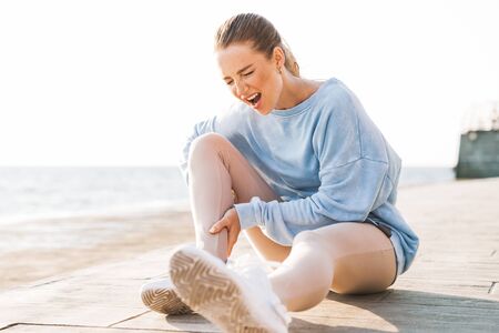 Picture of screaming young sports woman outdoors on beach touching leg feeling pain in calves.