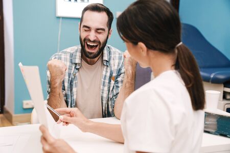 Image of female doctor in medical uniform and joyful patient man looking at health results in hospital roomの写真素材