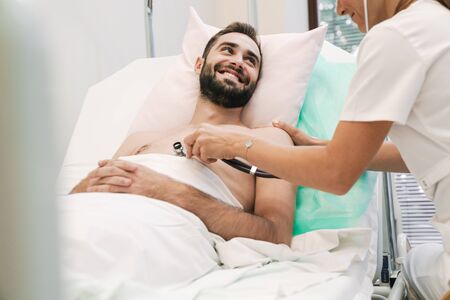 Image of female doctor in medical uniform examining young patient man in hospital bedの写真素材