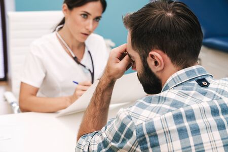 Image of concentrated serious doctor talking with her stressed sad patient in hospital at the table.