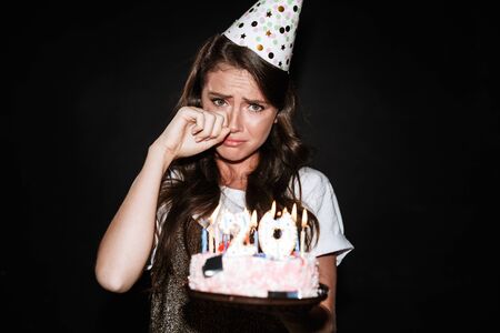 Image of beautiful upset woman in party cone holding cake with candles and crying isolated over black wallの写真素材