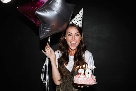 Image of young excited woman in party cone holding balloons and cake while smiling isolated over...