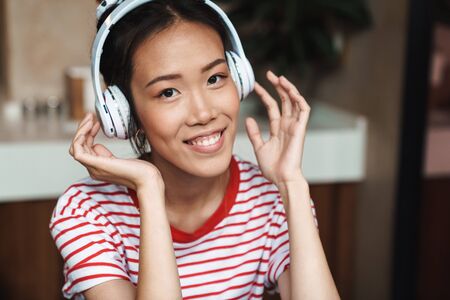 Portrait of joyful asian woman listening to music with headphones while sitting in cafe indoors
