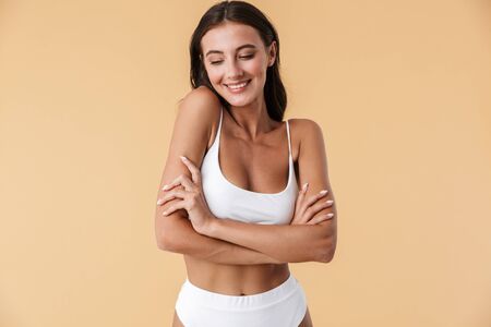 Portrait of a smiling beautiful sensual young brunette woman wearing lingerie isolated over beige background, posing, arms foldedの写真素材