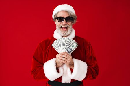 Portrait of excited old man wearing Santa Claus costume and sunglasses holding cash money isolated over red backgroundの写真素材