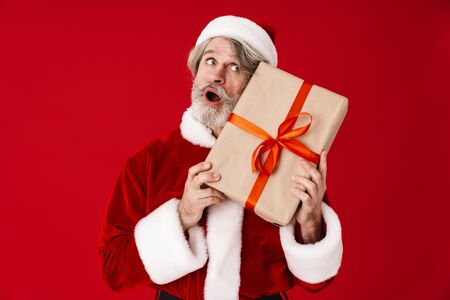 Image of joyful gray-haired old Santa Claus man laughing and holding gift box isolated over red background in studioの写真素材
