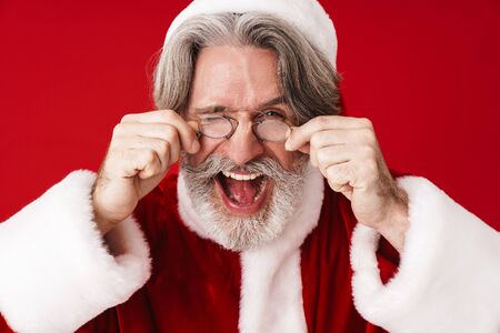 Image of optimistic gray-haired old Santa Claus man smiling and touching his spectacles isolated over red background in studioの写真素材