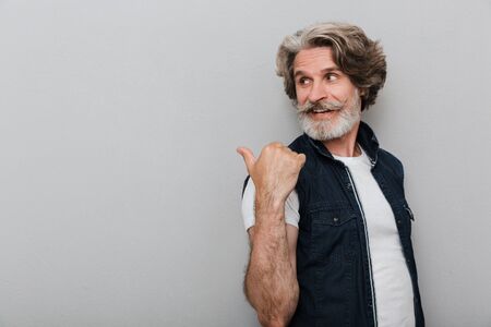 Image of a bearded cheery smiling senior man with grey hair isolated over grey wall background pointing aside.の写真素材