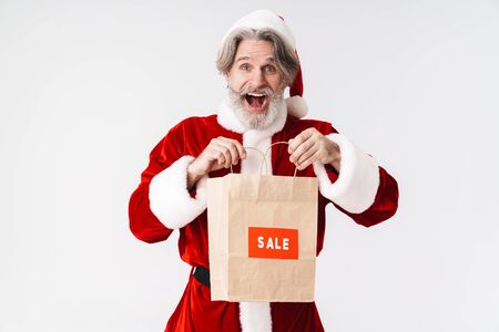 Portrait of delighted old man wearing Santa Claus red costume holding paper bag and screaming isolated over white backgroundの写真素材