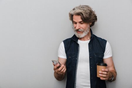 Portrait of a handsome smiling stylish mature man wearing a vest standing isolated over gray background, using mobile phone while drinking takeaway coffeeの写真素材