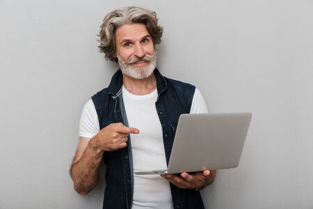 Portrait of a handsome smiling stylish mature man wearing a vest standing isolated over gray background, pointing at laptop computerの写真素材