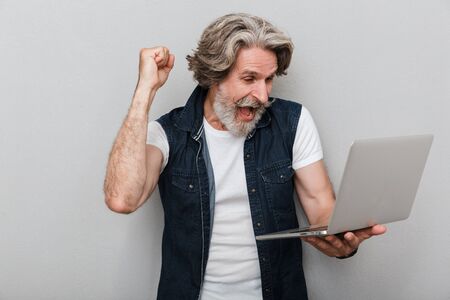 Portrait of a handsome stylish mature man wearing a vest standing isolated over gray background, using laptop computer, celebrating successの写真素材