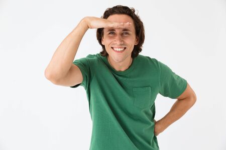 Image of optimistic young man wearing basic t-shirt smiling and watching with his hand at forehead...