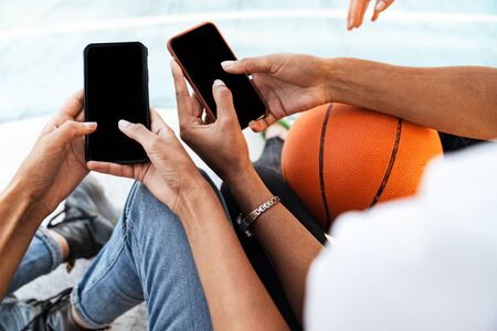 Cropped image of two young african american girls in trendy streetwear sitting on playdround with basketball and smartphonesの写真素材