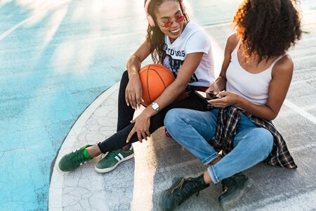 Image of two happy african american girls in trendy streetwear sitting on playdround with basketballの写真素材