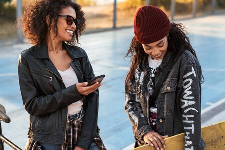 Image of two happy african american girls in trendy streetwear talking and holding smartphone outdoorsの写真素材