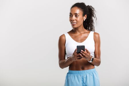 Portrait of smiling african american woman in sportswear using cellphone and earpods isolated over white backgroundの写真素材