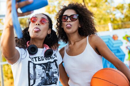 Image of two pretty african american girls in casual clothes taking selfie on hipster photo camera...
