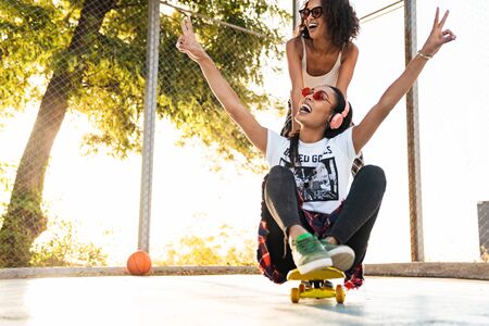 Image of two excited african american girls having fun and riding skateboard together on playgroundの写真素材