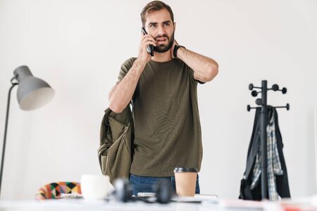 Image of handsome perplexed man with bag talking on cellphone and touching his neck while working in bright officeの写真素材